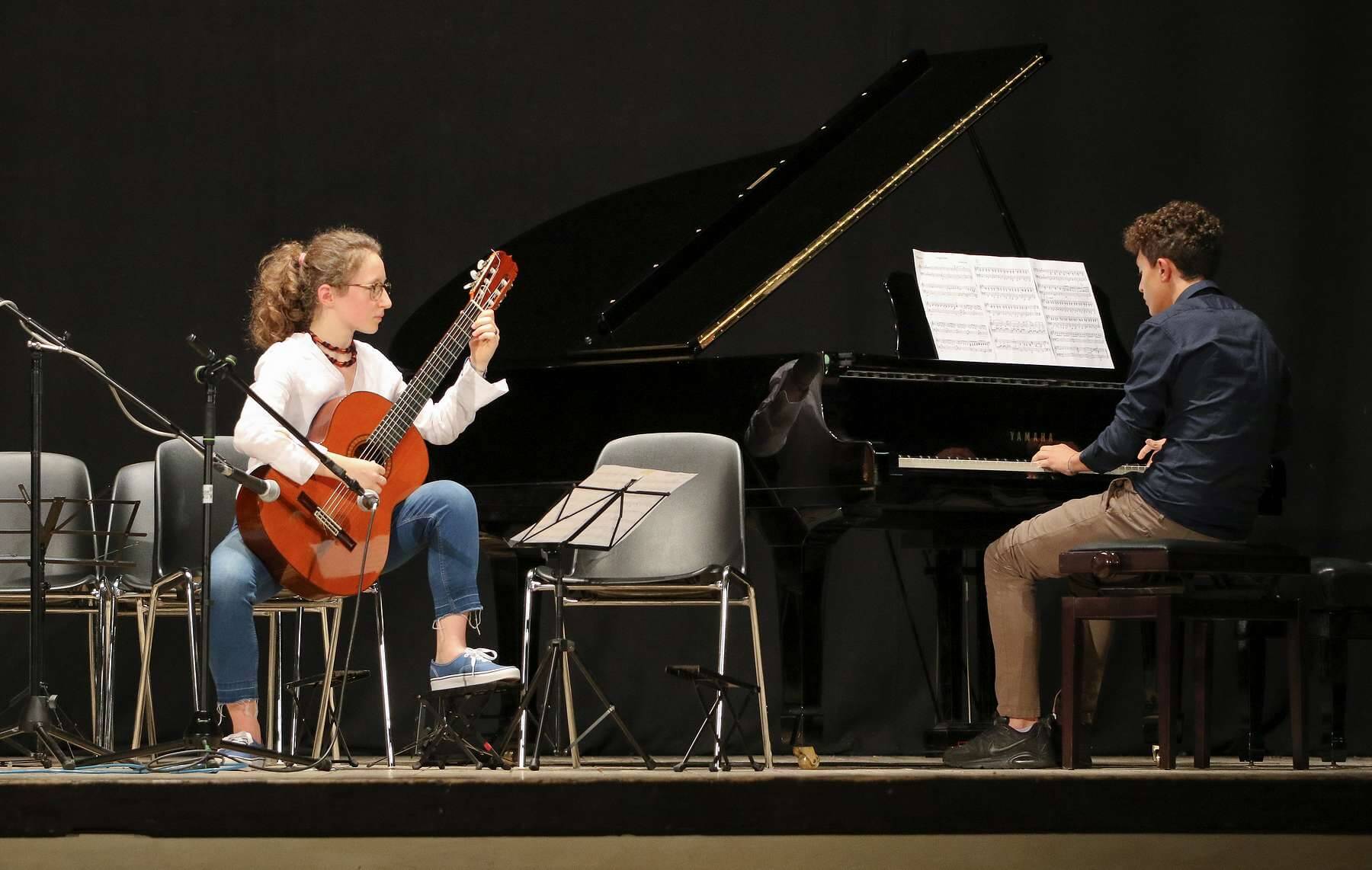 Una ragazza suona la chitarra accompagnata da un ragazzo che suona il pianoforte durante un concerto